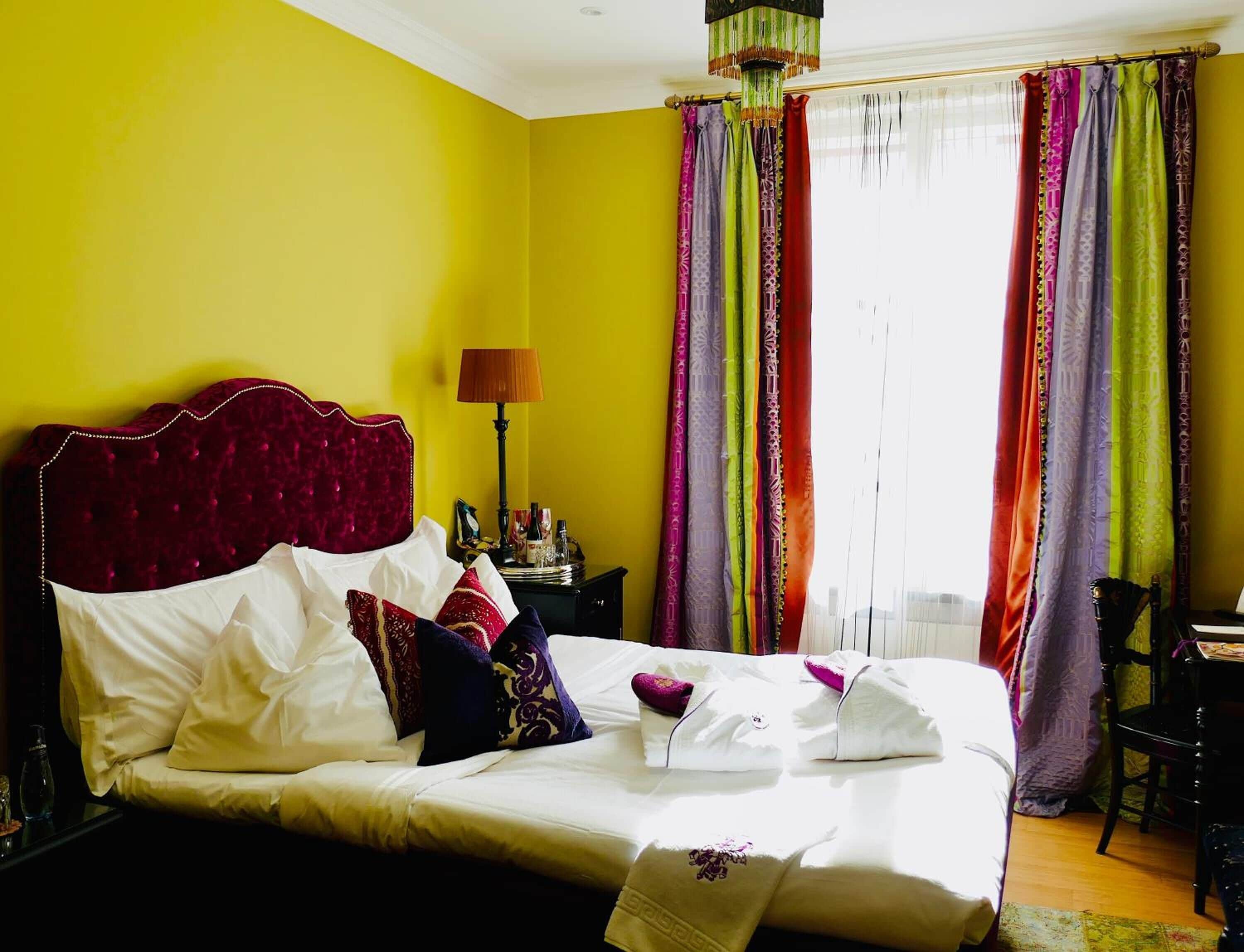 A bed with white linens and a red headboard in a room with yellow walls and colorful curtains.