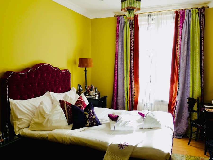 A bed with white linens and a red headboard in a room with yellow walls and colorful curtains.