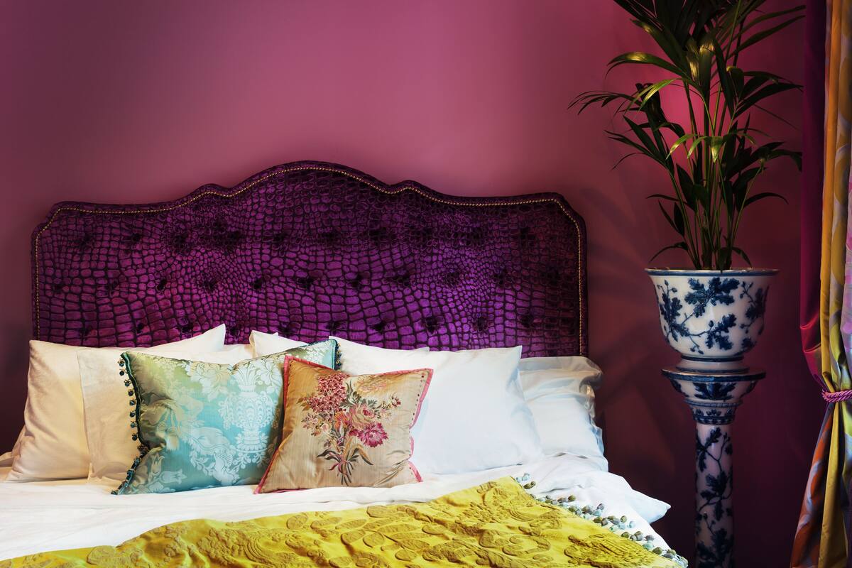 Un lit avec plusieurs oreillers et une couverture jaune, un grand vase avec une plante et une tête de lit à motifs.