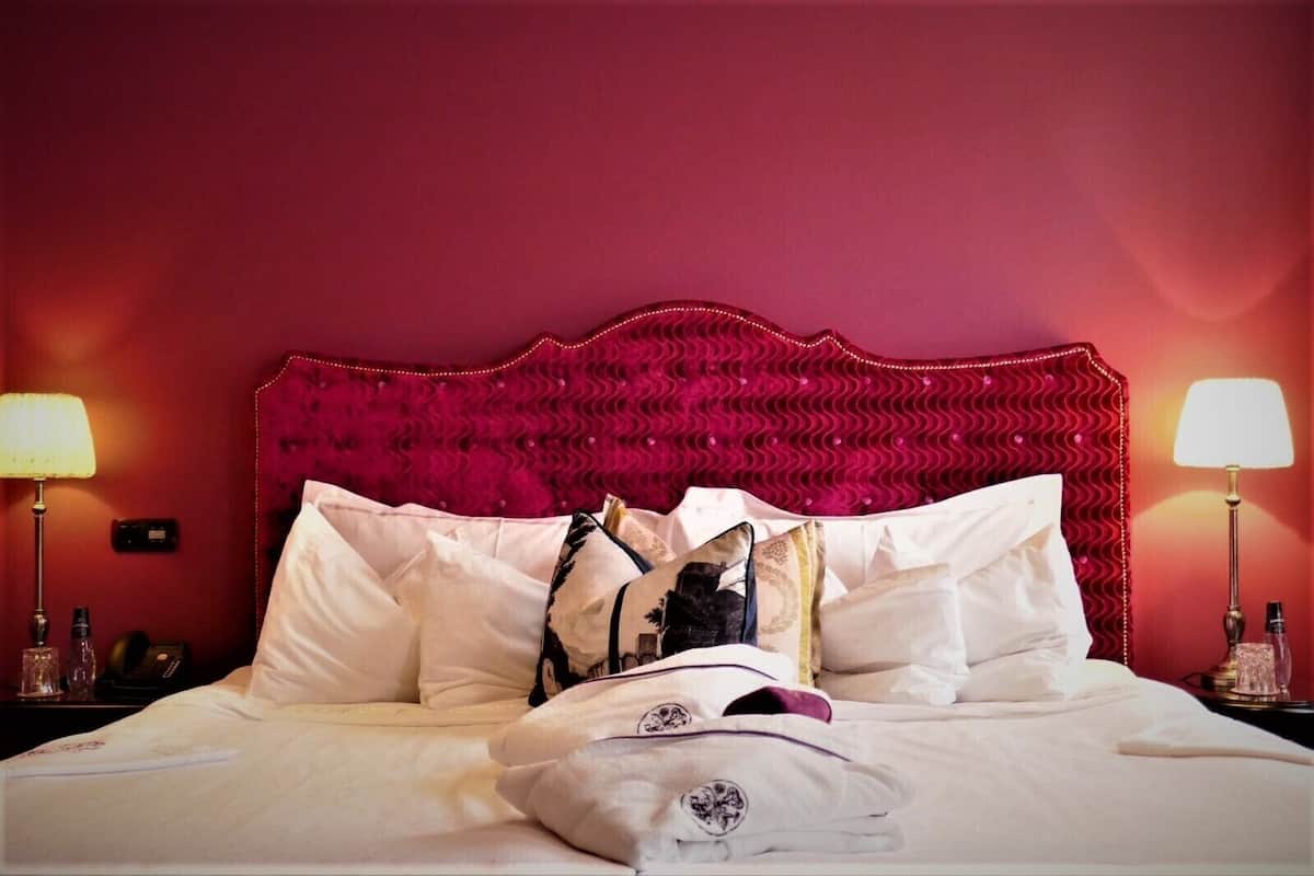Une chambre d’hôtel avec une tête de lit rembourrée de tissu rouge, du linge de lit blanc et deux lampes de chevet.