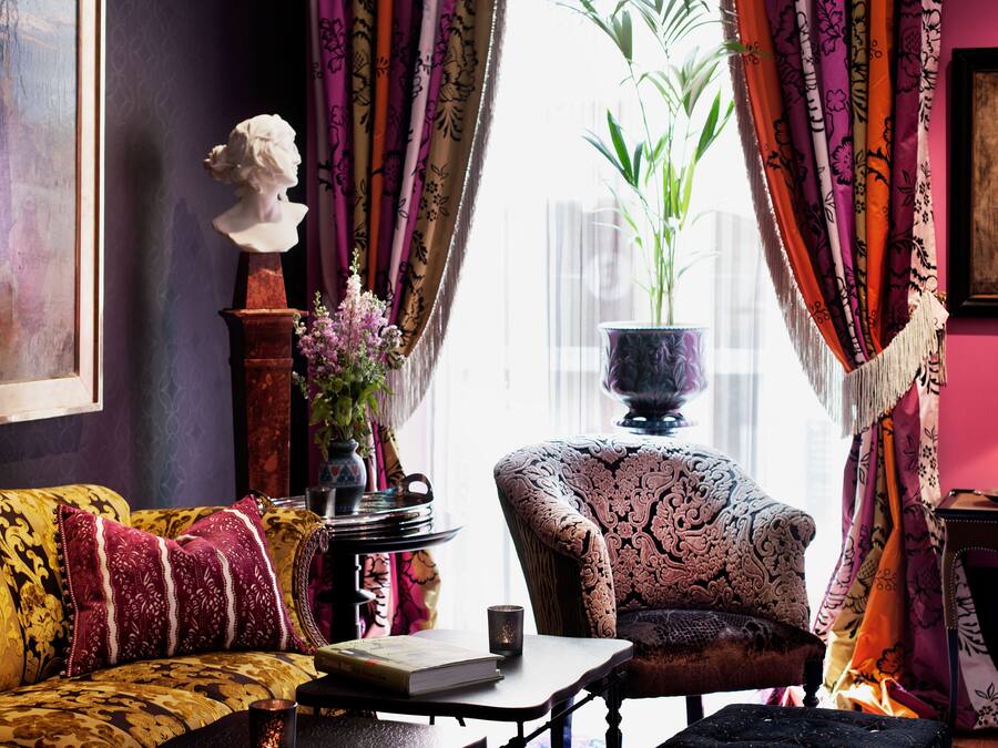 Una habitación con un sofá estampado, una silla, una mesa con un libro y una taza, un jarrón con flores y un cuadro en la pared.