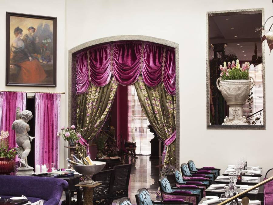 Un comedor con una larga mesa dispuesta para una comida, flanqueado por cortinas moradas y verdes, y una estatua de una mujer con un vestido rojo.
