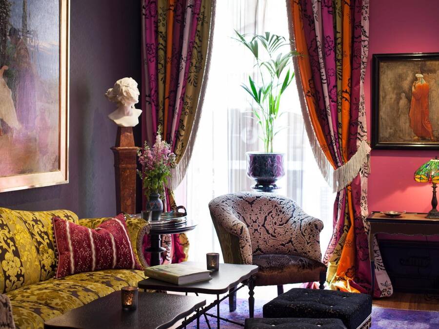 A room with a patterned sofa, a small table with a book and a candle, a chair, a potted plant, and a painting on the wall.