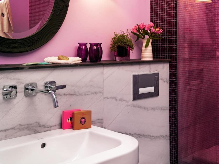 Un baño con lavamanos blanco, inodoro y espejo. Hay dos jarrones morados con flores en la pared, y una planta pequeña en maceta.