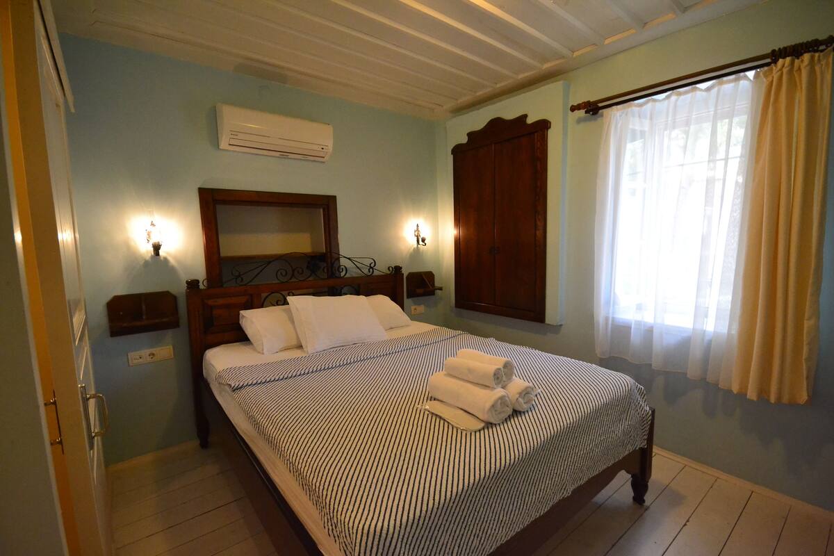 A bedroom with a bed, bedside lamps, an air conditioner, a window with curtains, and a wooden wardrobe.