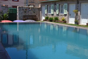 A swimming pool with a stone wall and shutters, surrounded by potted plants and a bench.
