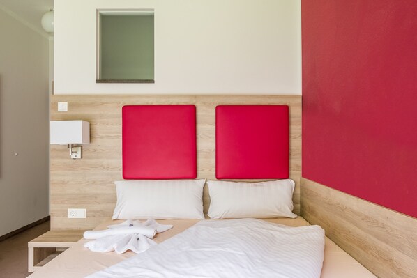 Habitación de hotel con cabecera de madera, dos almohadas rojas, cubrecama blanco y toalla blanca sobre la cama.