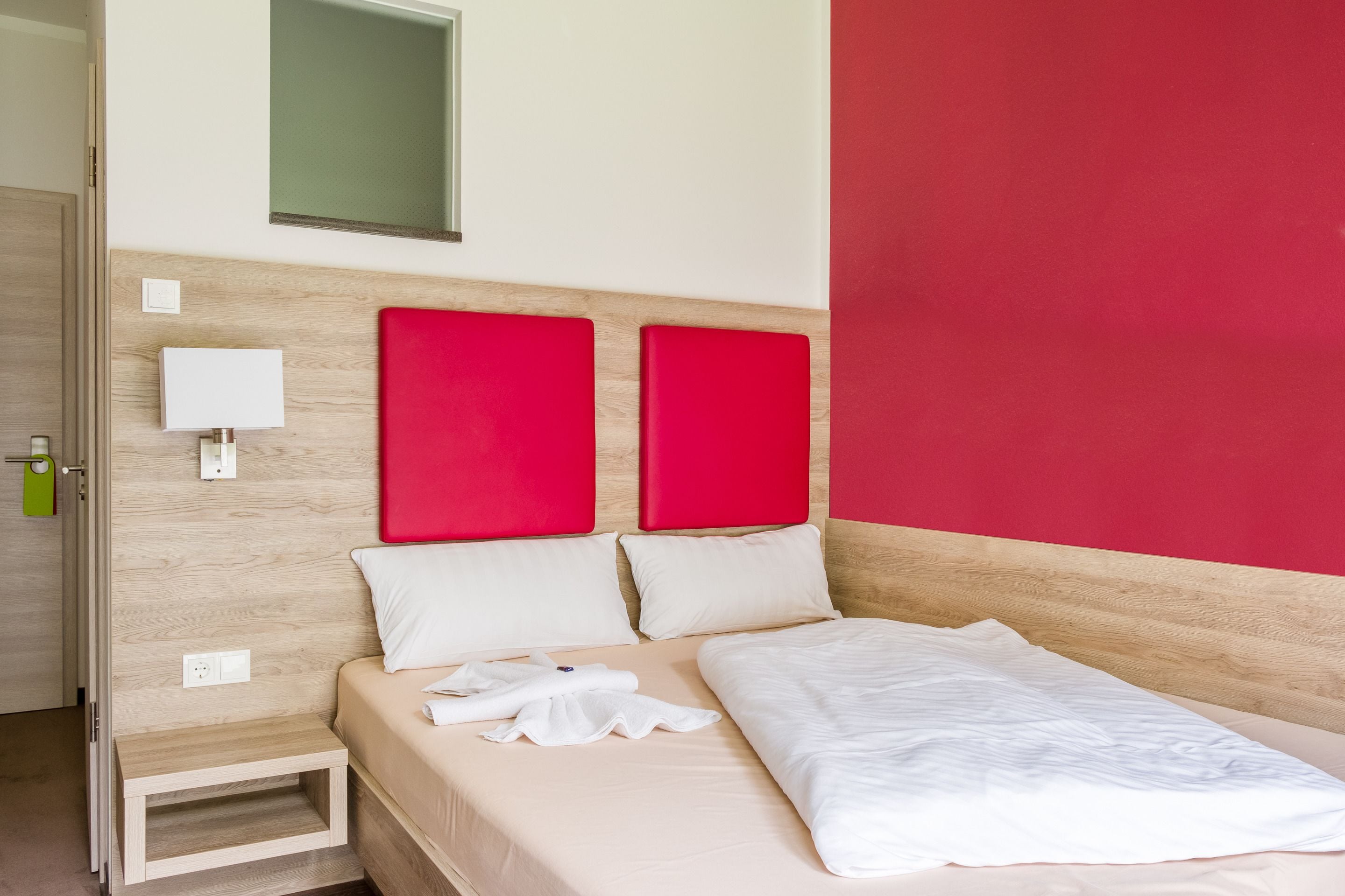 A neatly made bed with white linens, a small bedside table, and a red accent wall.