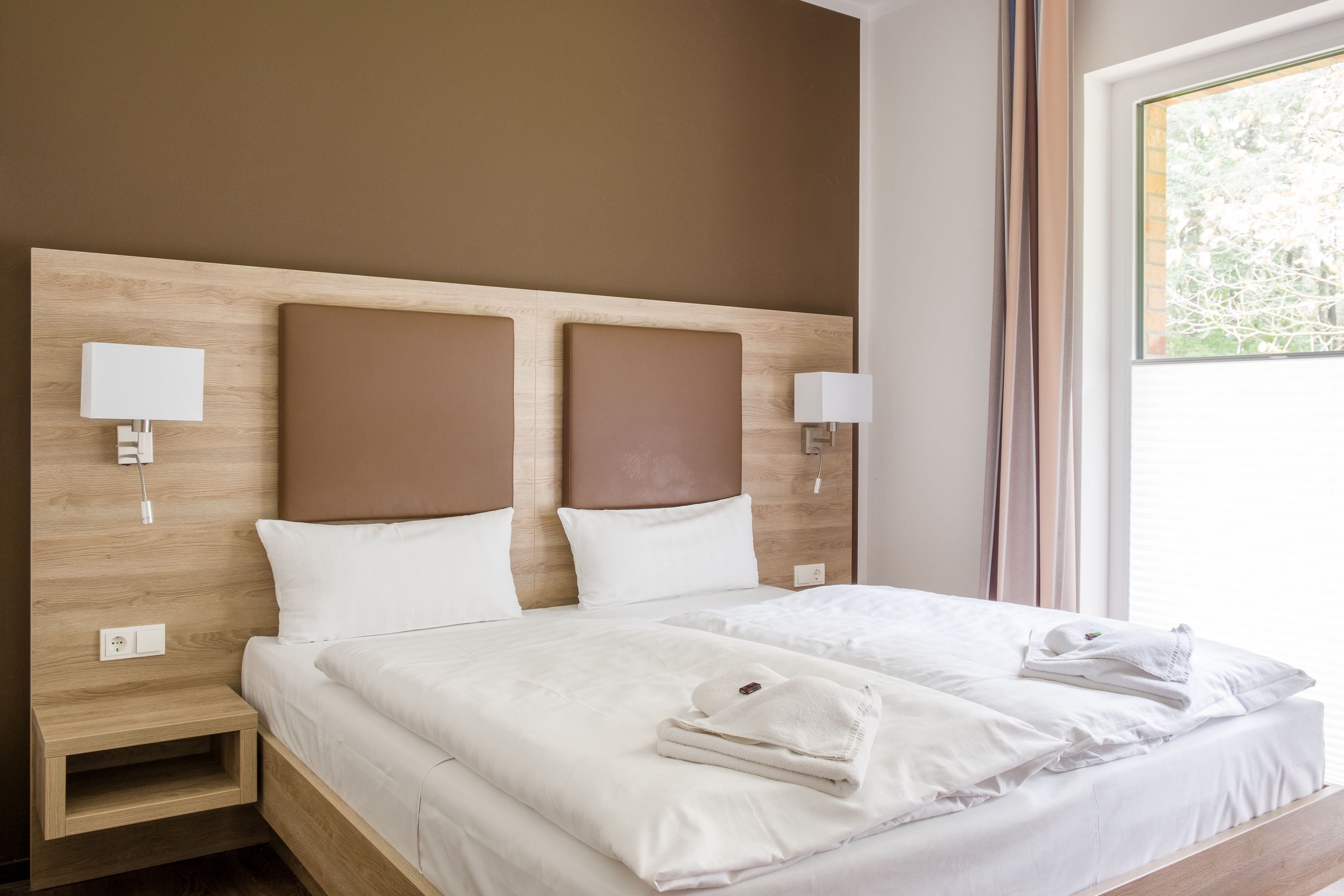 Una cama bien hecha con sábanas blancas y una toalla doblada al lado. Hay dos mesitas de noche con lámparas y un enchufe en la pared.