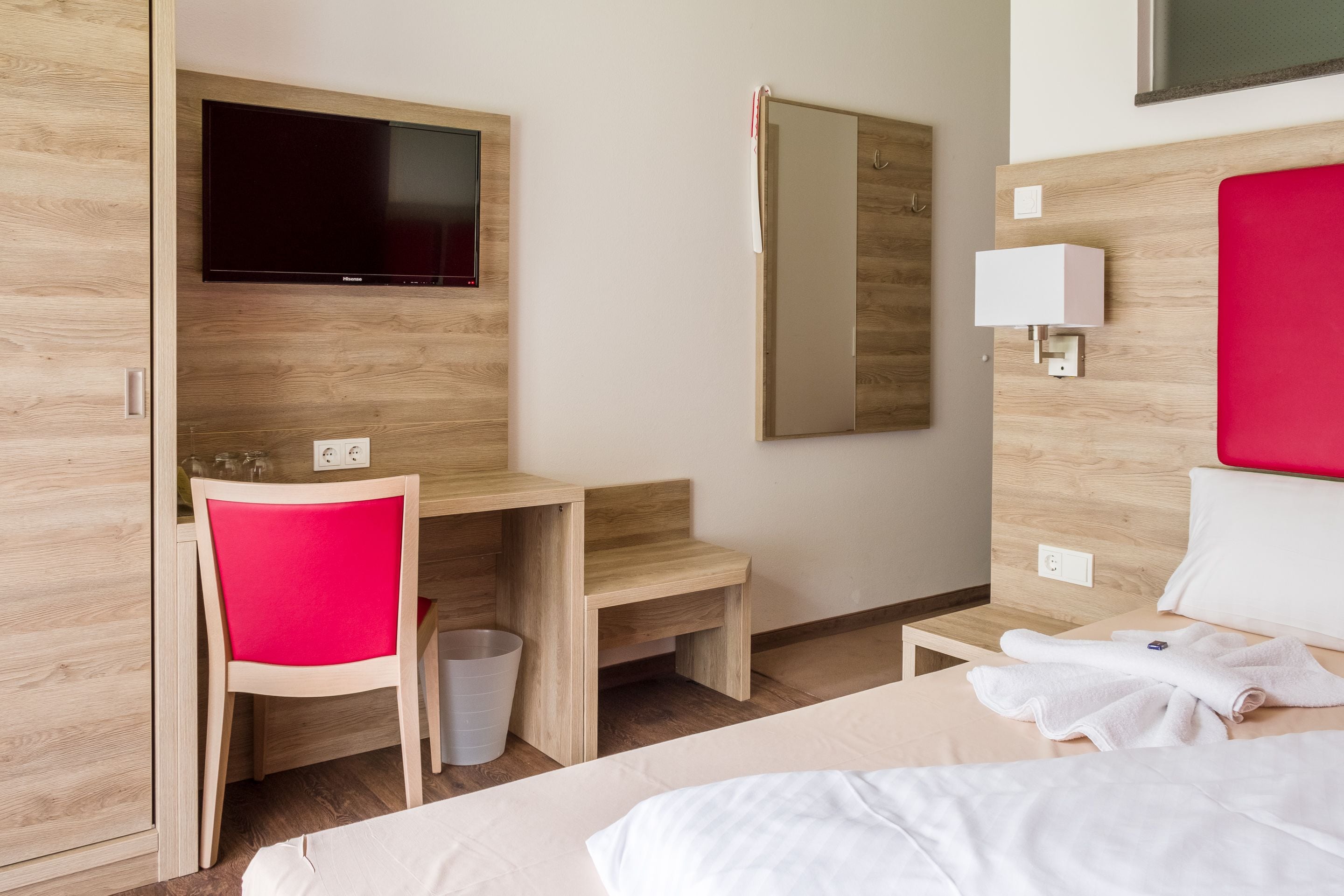 A hotel room with a bed, a desk with a chair, a television, a mirror, and a bedside table with a lamp.
