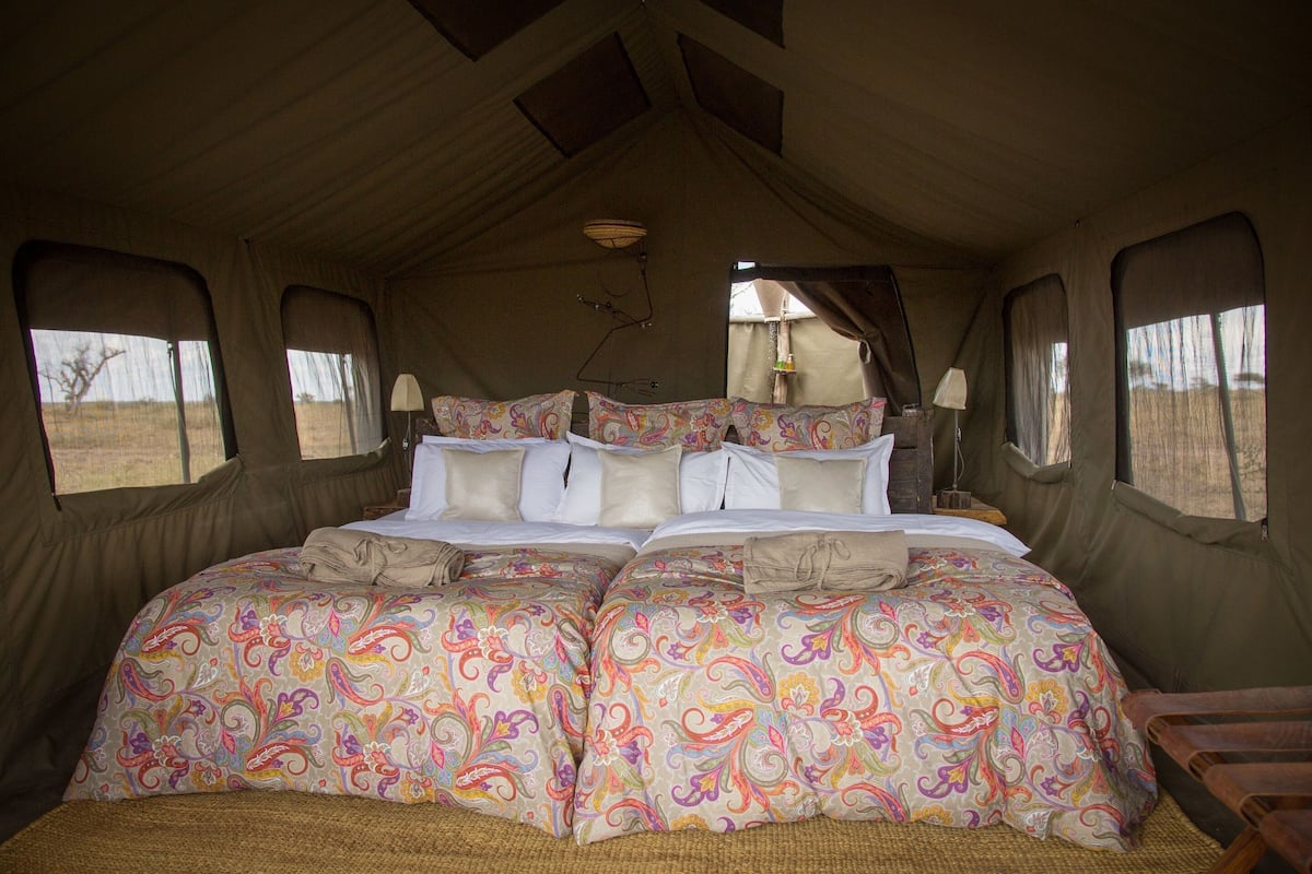 A tented accommodation with a bed featuring patterned bedding, a lamp, and a view of the outdoors.