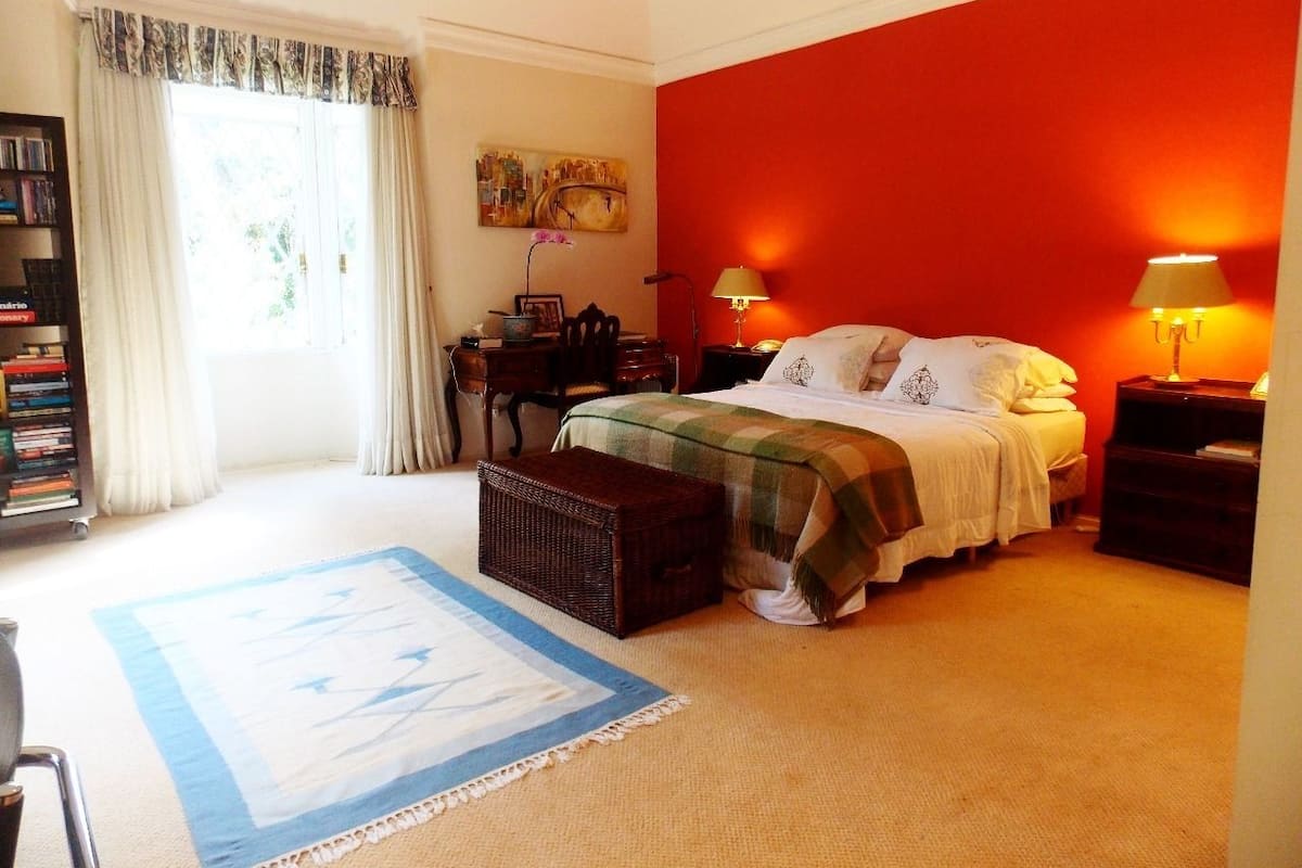 A bedroom with a bed, bedside tables, a nightstand, a bookshelf, a window with curtains, and a painting on the wall.