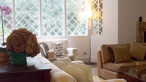 A living room with a sofa, armchairs, a coffee table, and a lamp. There is a window with a lattice design and a vase with flowers on a table. - Casa Yvitú Blue Bird Hotel (Campos do Jordão)