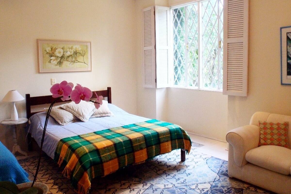 A bedroom with a bed, a chair, a window with shutters, and a painting on the wall.
