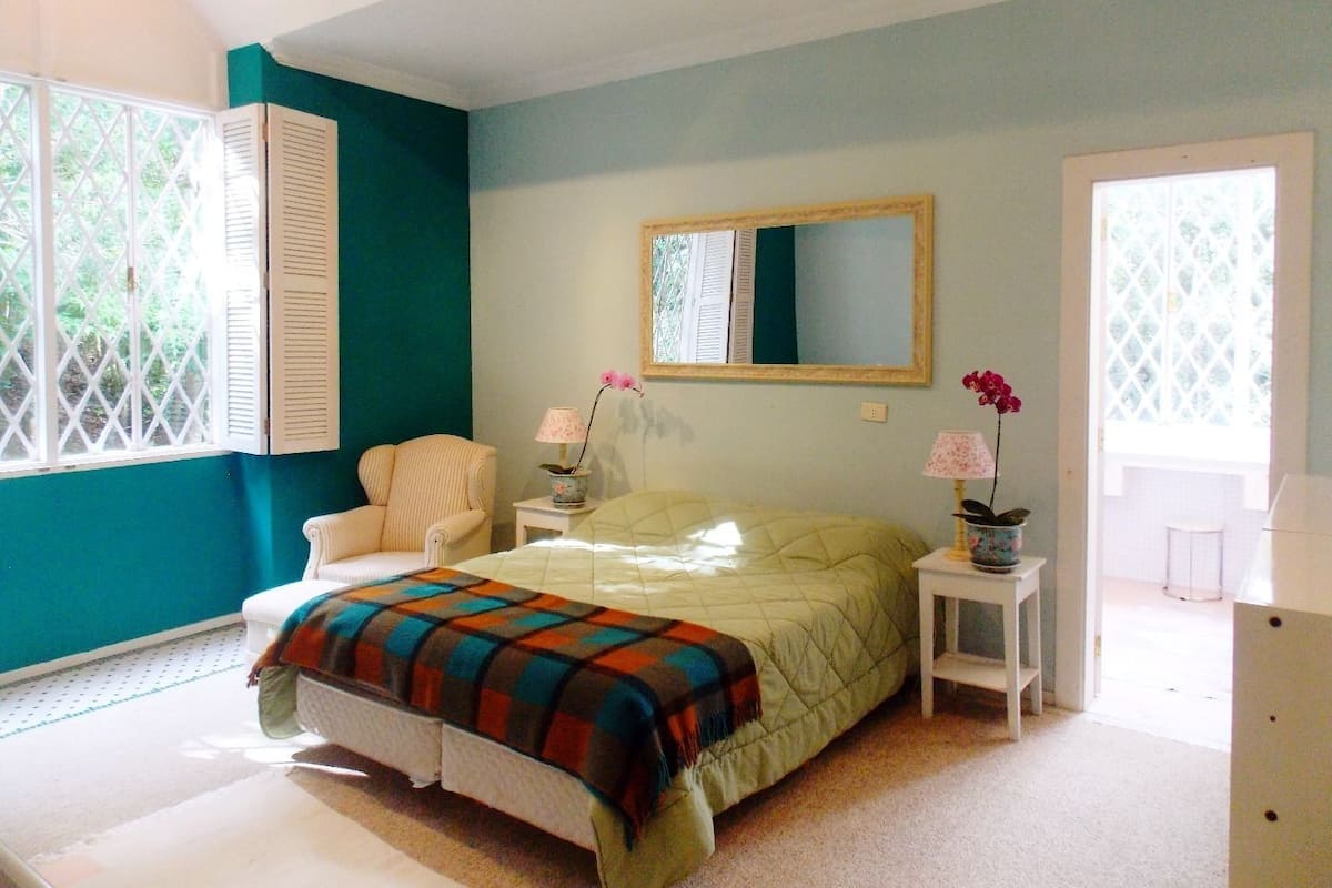 A bedroom with a bed, a chair, a nightstand, a mirror, and a window with shutters.