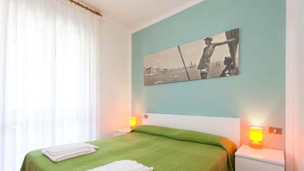 A hotel room with a bed, green bedspread, white towels, a painting on the wall, and two bedside lamps. - Villa Valentina (Jesolo)
