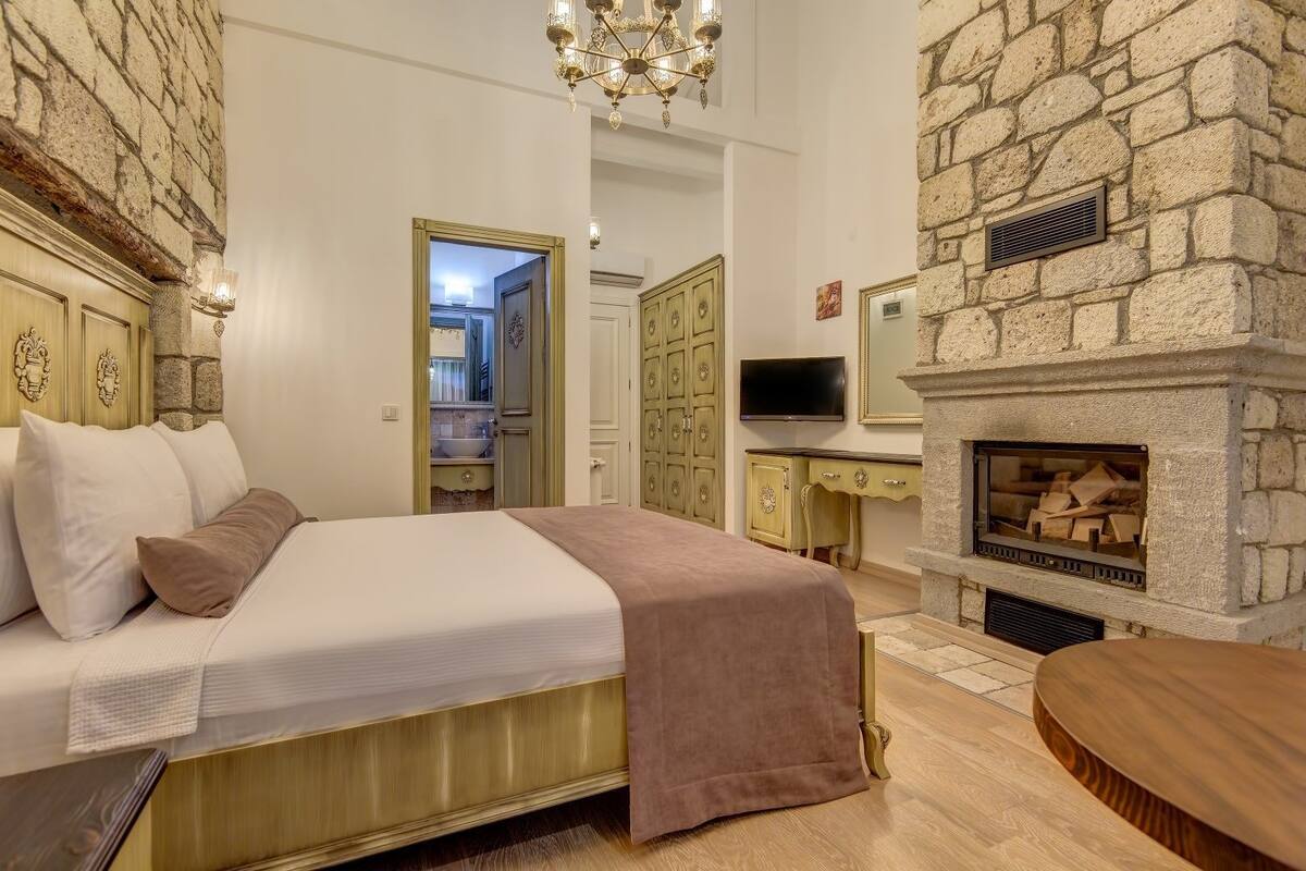 Un dormitorio con cama, chimenea de piedra, televisión y un baño visible a través de una puerta abierta.