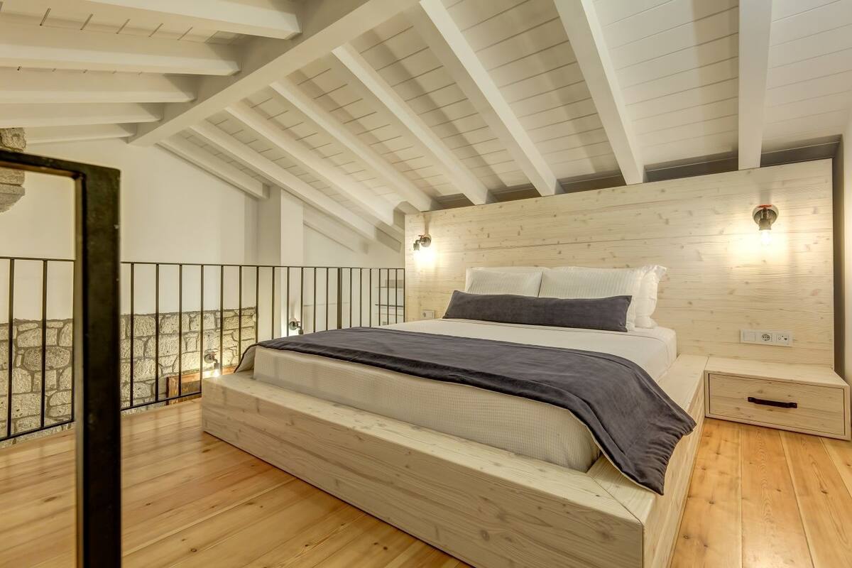 Un dormitorio con cabecera de madera, cama con ropa de cama blanca y gris, mesita de noche y suelo de madera.