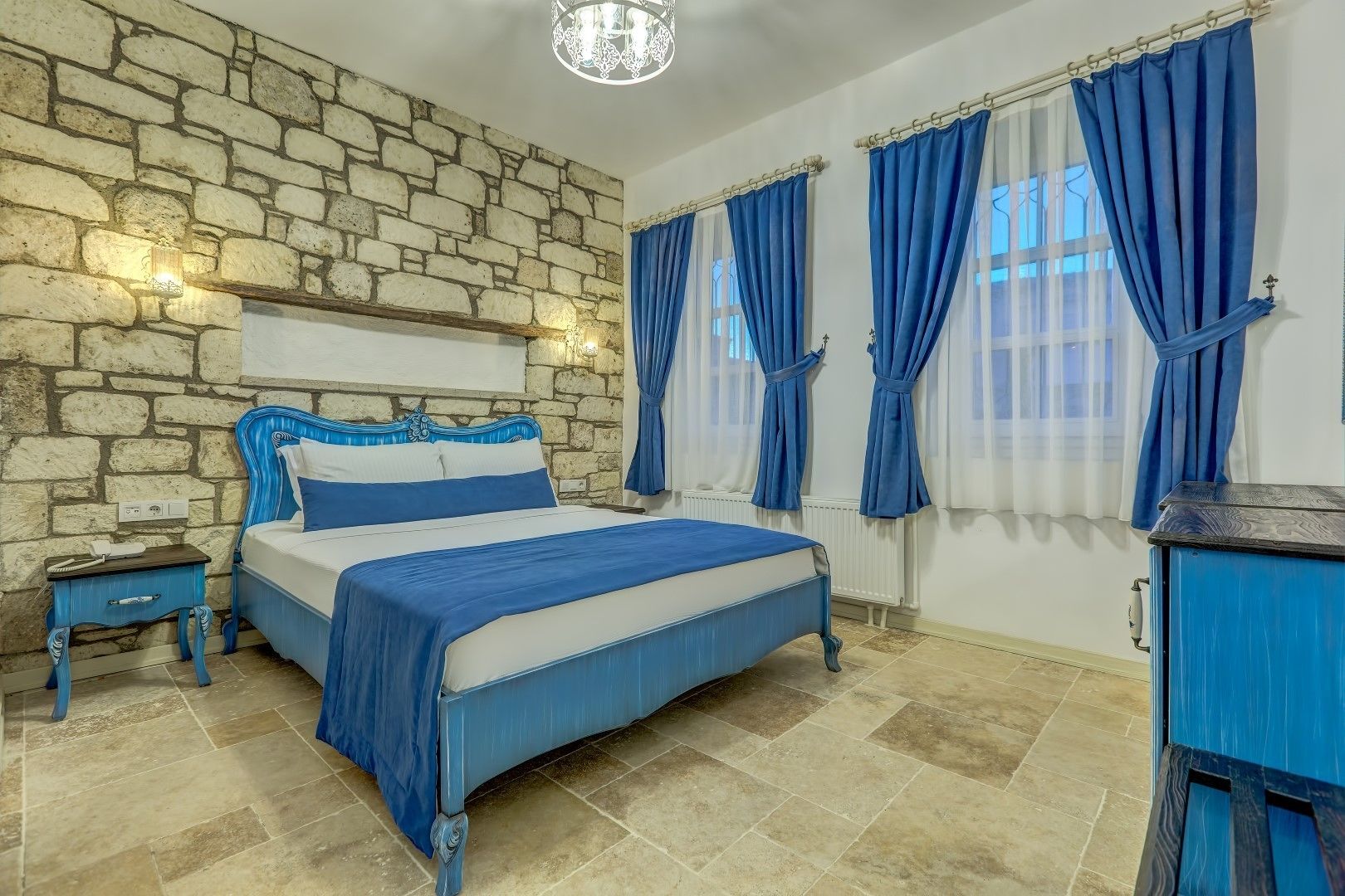 Un dormitorio con pared de piedra, cama con ropa de cama azul, mesita de noche y cómoda.