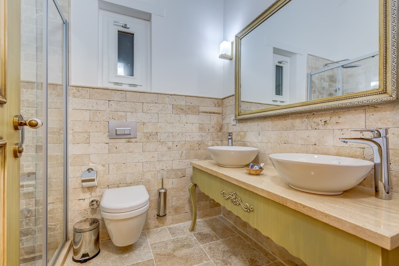 Ein Badezimmer mit Duschkabine aus Glas, zwei Waschbecken, einer Toilette und einem Spiegel mit Zierrahmen.