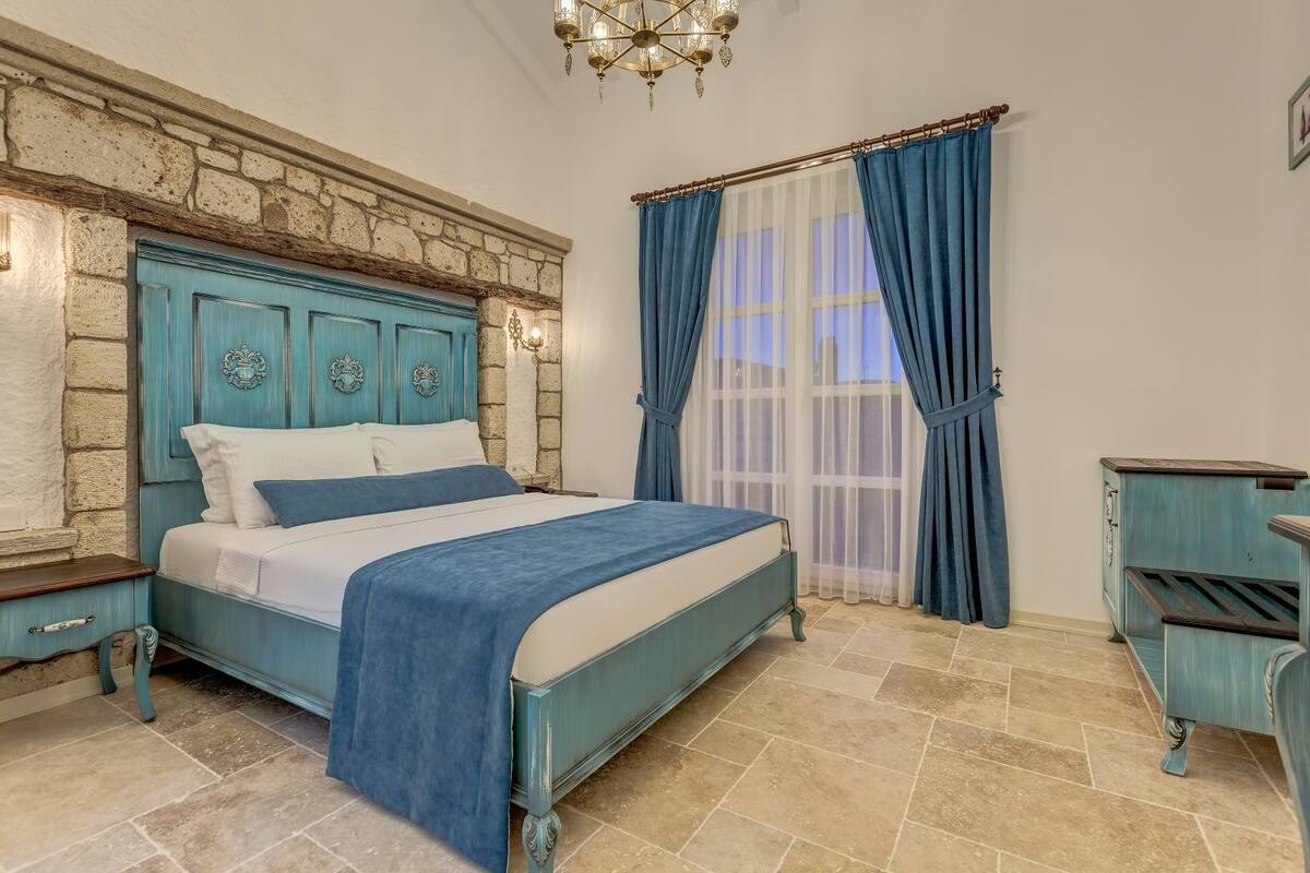 Un dormitorio con pared de piedra, una cama con cabecera azul, una mesita de noche, un aparador y una araña.