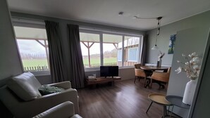 Una sala de estar con un sofá, una mesa de centro y un televisor. Hay ventanas grandes con cortinas, un comedor con sillas y un jarrón blanco con flores en la pared.