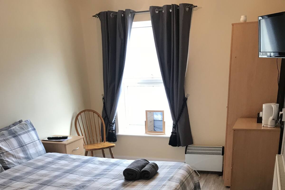 Quarto com cama, cadeira, mesinha, radiador e janela com cortinas.