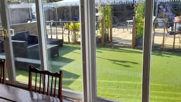 Un área de comedor con una mesa y sillas, puertas de cristal correderas grandes que dan a un jardín con césped y una estructura de invernadero.