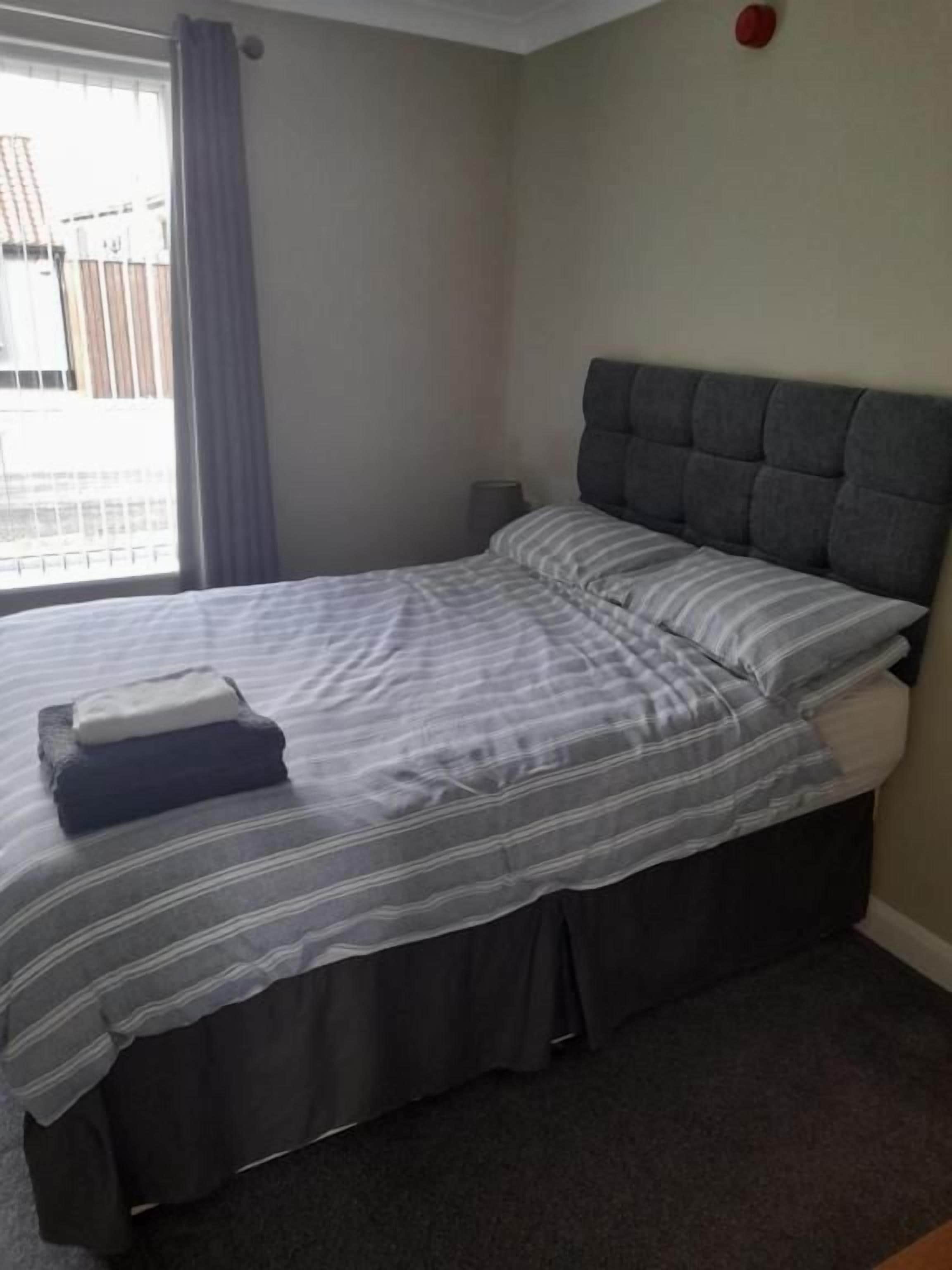 Un dormitorio con cama, cabecera, ventana con cortinas y una mesita de noche.