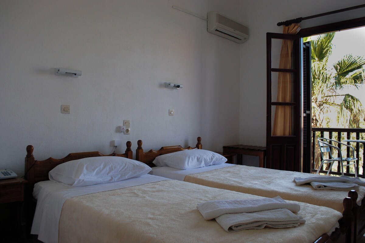 A room with two beds, a wooden headboard, a small table, and a balcony with a chair.