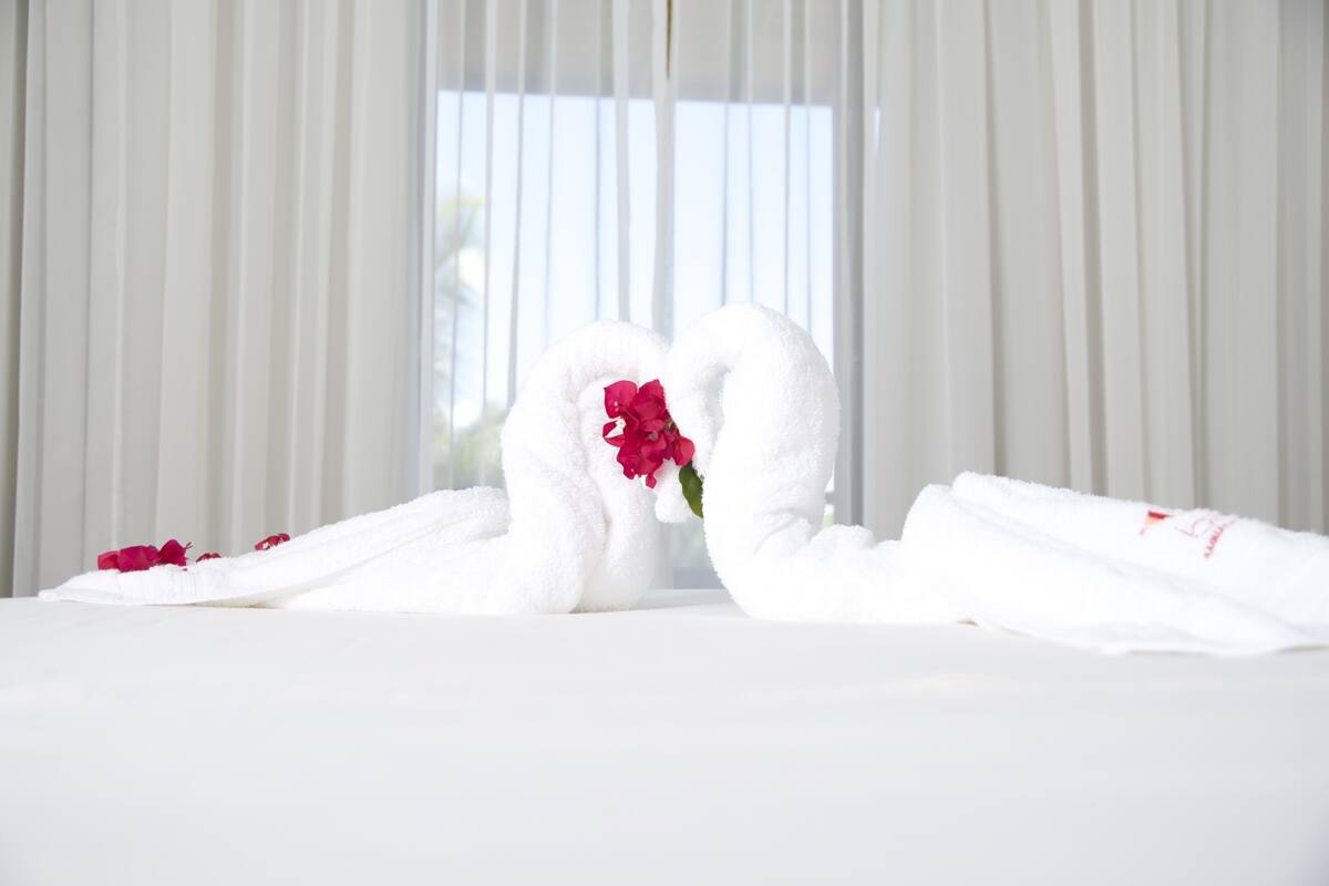A bed with white linens and two towels arranged in a heart shape, with a red flower in the center.