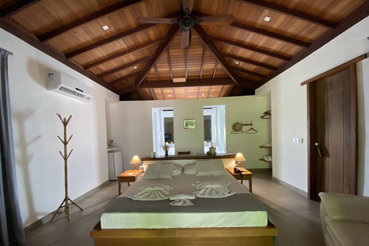 A bedroom with a wooden ceiling, a bed with white bedding, bedside lamps, a coat rack, and a small desk.