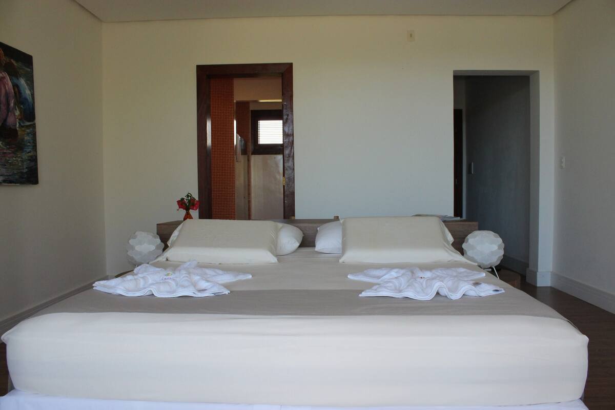 A double bed with white linens and pillows, a vase with flowers, and a painting on the wall.