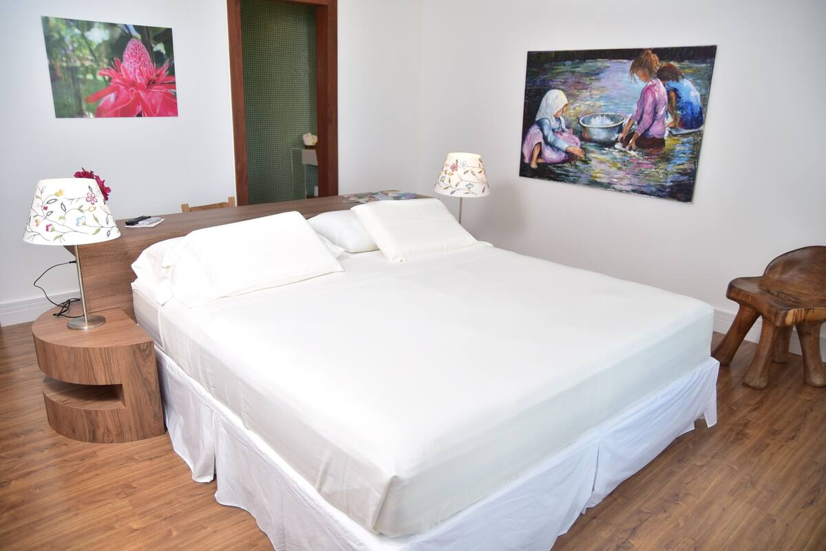 A bedroom with a large bed, two bedside lamps, a wooden nightstand, a wooden chair, and two paintings on the wall.