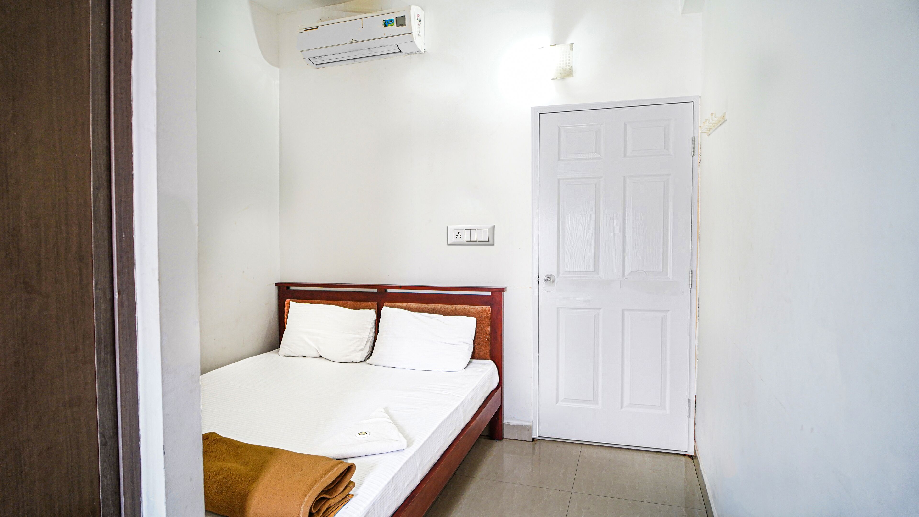 A bedroom with a wooden bed, white bedding, a white door, and an air conditioning unit on the wall.