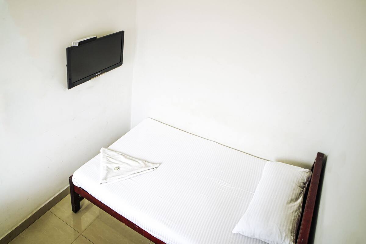 A small, single-bed room with a wooden frame, white bedding, and a flat-screen TV mounted on the wall.