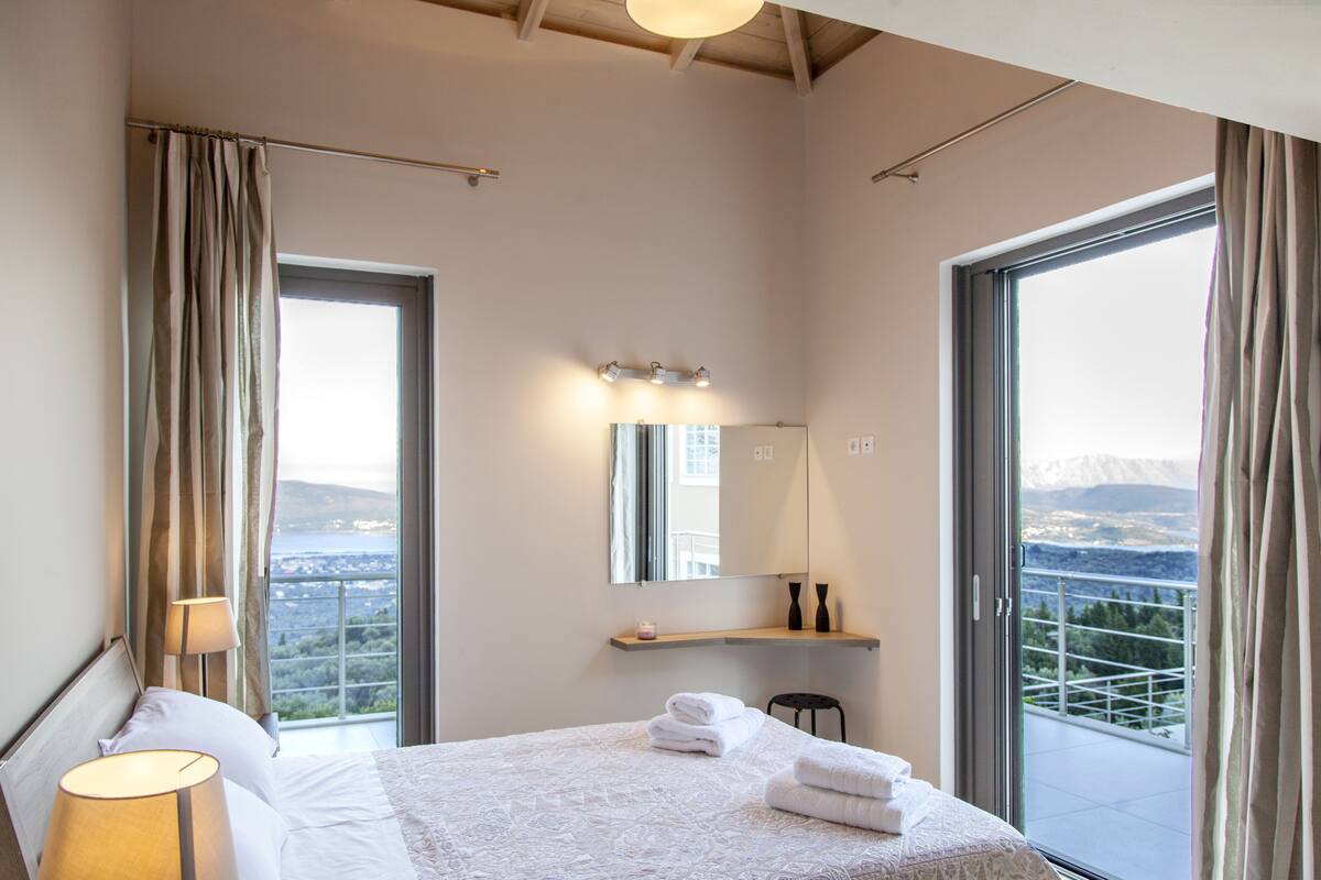 A modern hotel room with a large bed, a sitting area, a balcony with a view, and a bathroom with a mirror and lights.