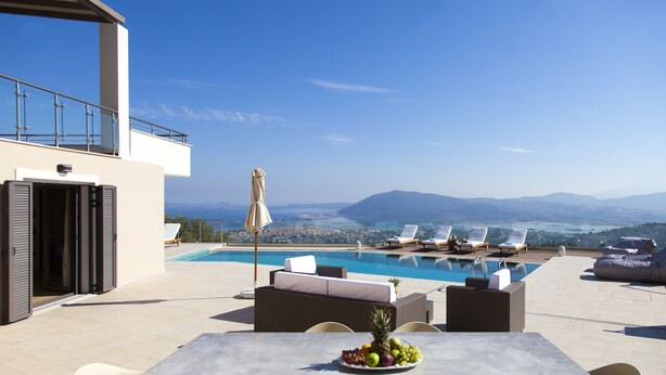 A rooftop pool area with a table set with fruit, lounge chairs, and a view of the city and mountains. - Alivia Lefkas (Lefkada)