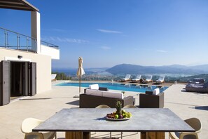 A rooftop pool area with a table set with fruit, lounge chairs, and a view of the city and mountains. - Alivia Lefkas (Lefkada)