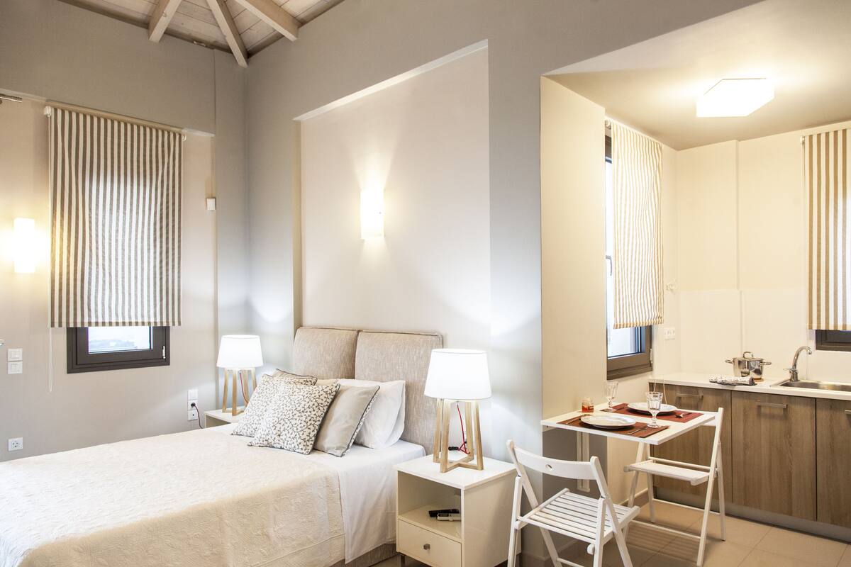 A modern hotel room with a bed, bedside tables, a desk, and a kitchenette.