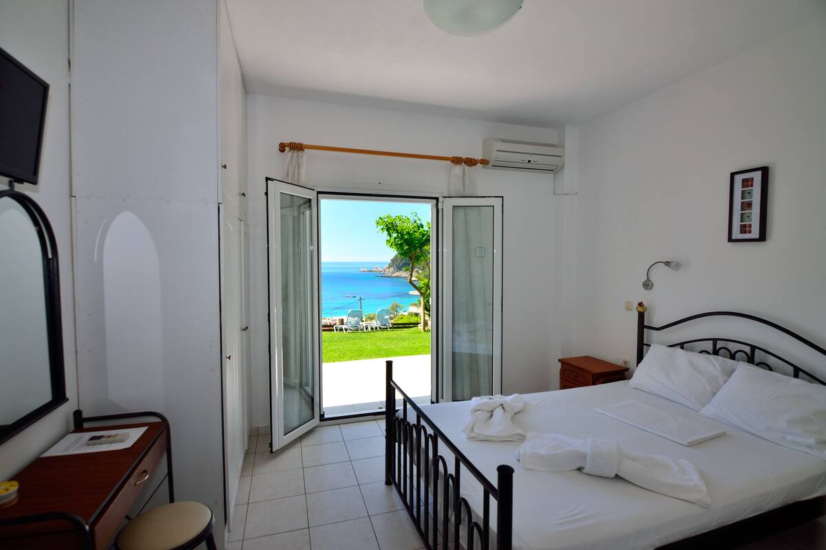 A bedroom with a bed, a desk, a chair, a TV, and a view of the sea.