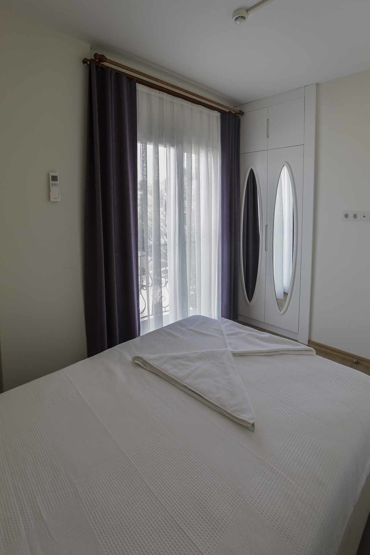 A hotel room with a bed, a window with curtains, a wardrobe, and a wall-mounted air conditioning unit.