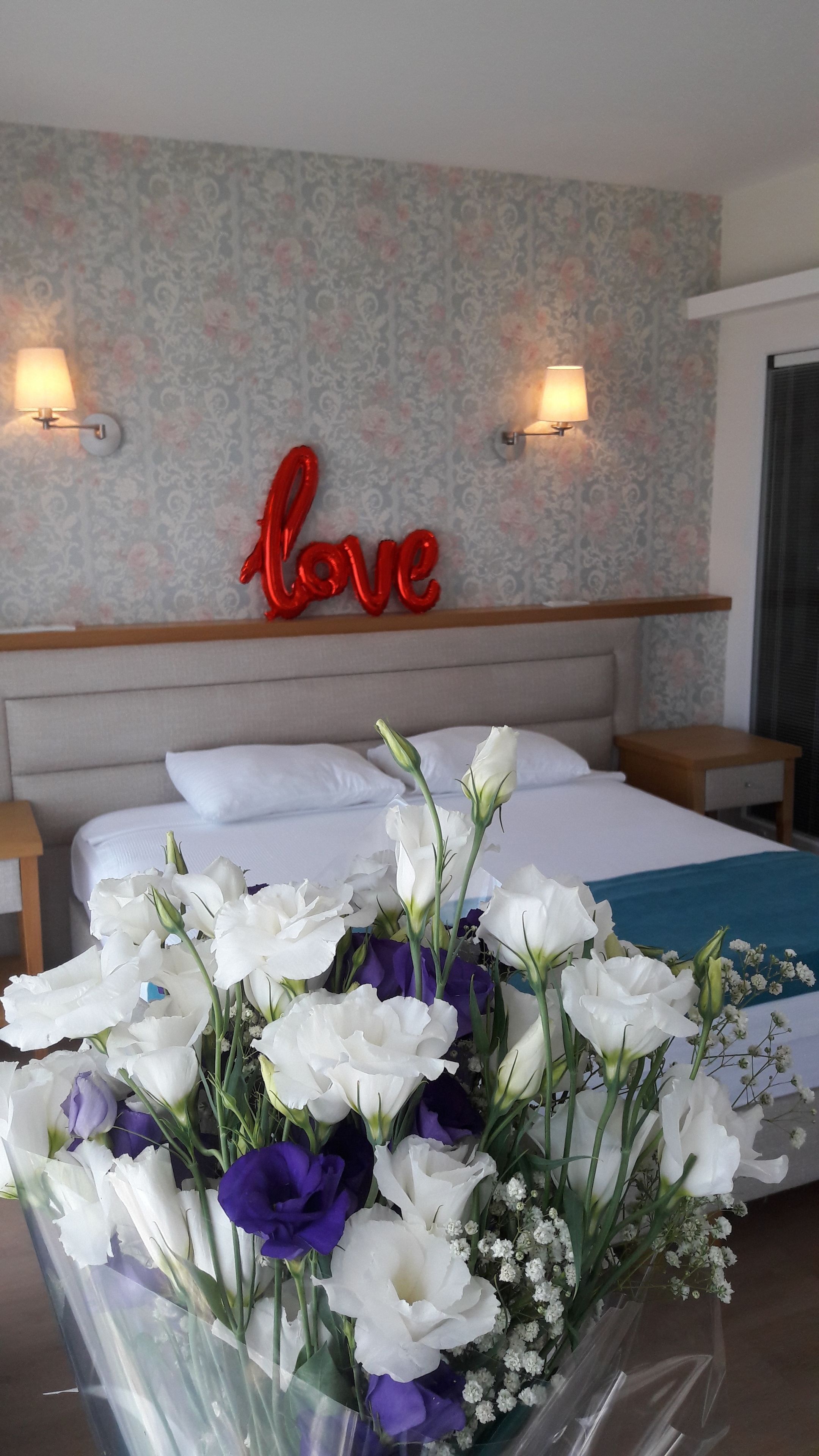 Een hotelkamer met een bed, een muur met een 'love'-bord en een bloemenboeket op het nachtkastje.