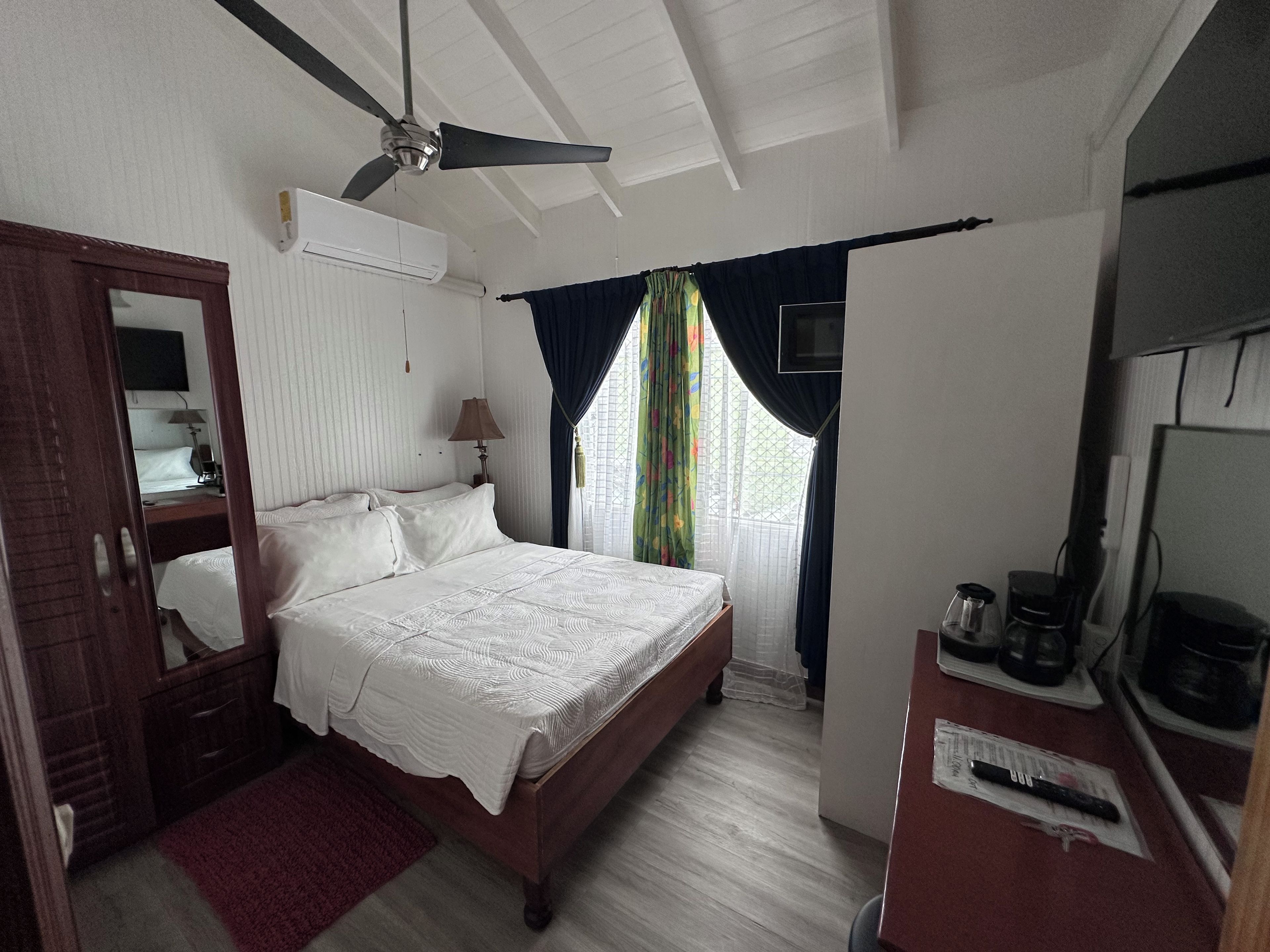 Un dormitorio con cama, ventilador de techo, televisión, escritorio y cafetera, y ventana con cortinas.