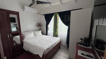 Un dormitorio con cama, ventilador de techo, televisión, escritorio y cafetera, y ventana con cortinas.