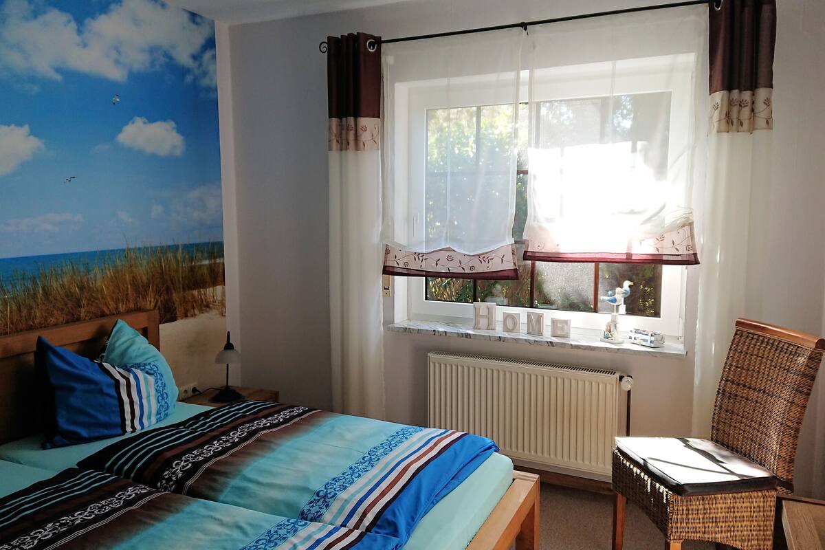 Ein Schlafzimmer mit einem Bett, einem Sessel, einem Fenster mit Vorhängen und Blick auf den Strand.