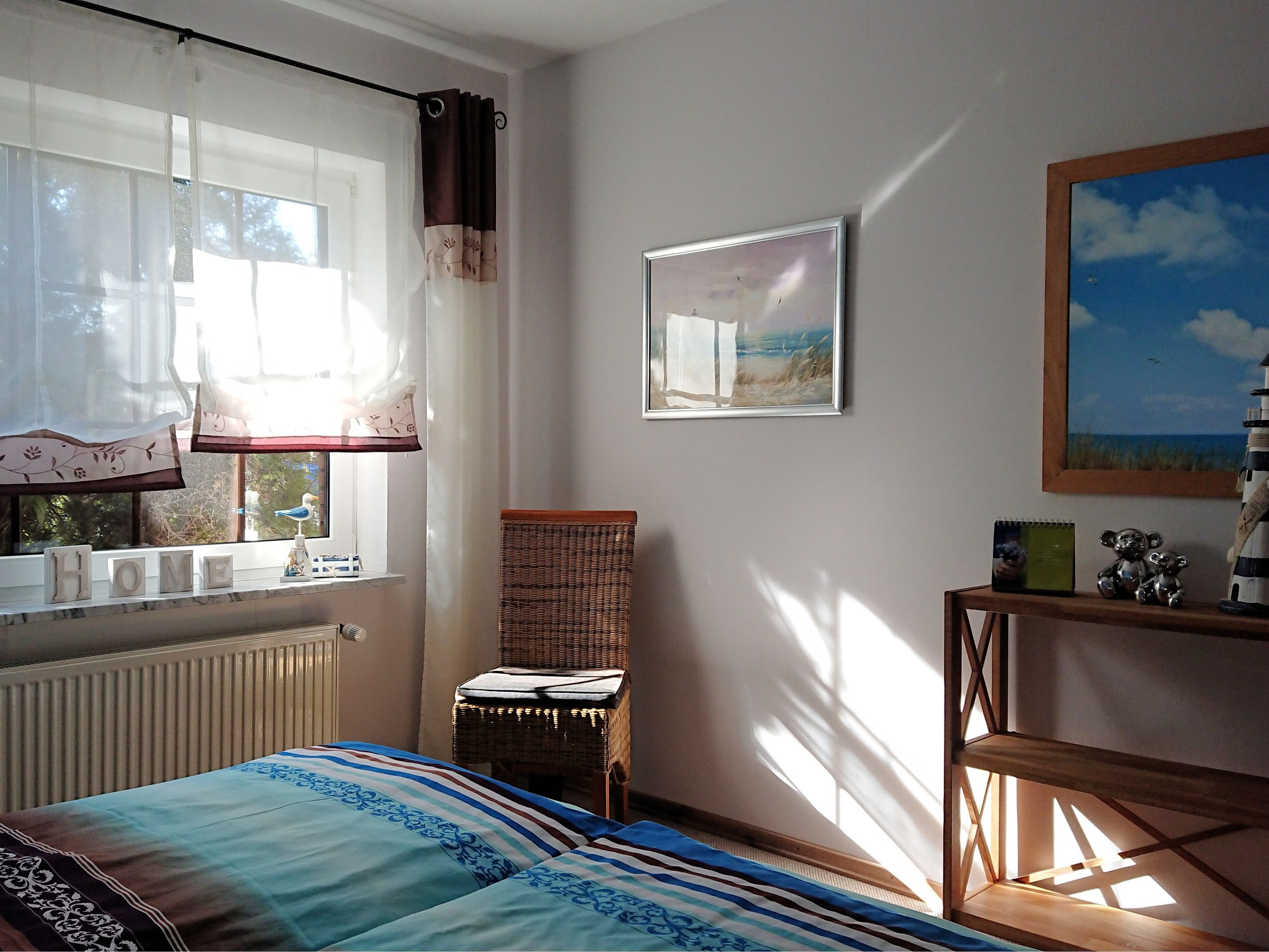 A bedroom with a bed, a chair, a window with curtains, a radiator, a shelf with items, and a painting on the wall.