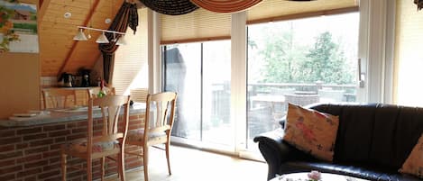 A cozy living room with a wooden ceiling, a black leather couch, a dining area with wooden chairs, and a kitchen area with a brick wall and wooden cabinets.