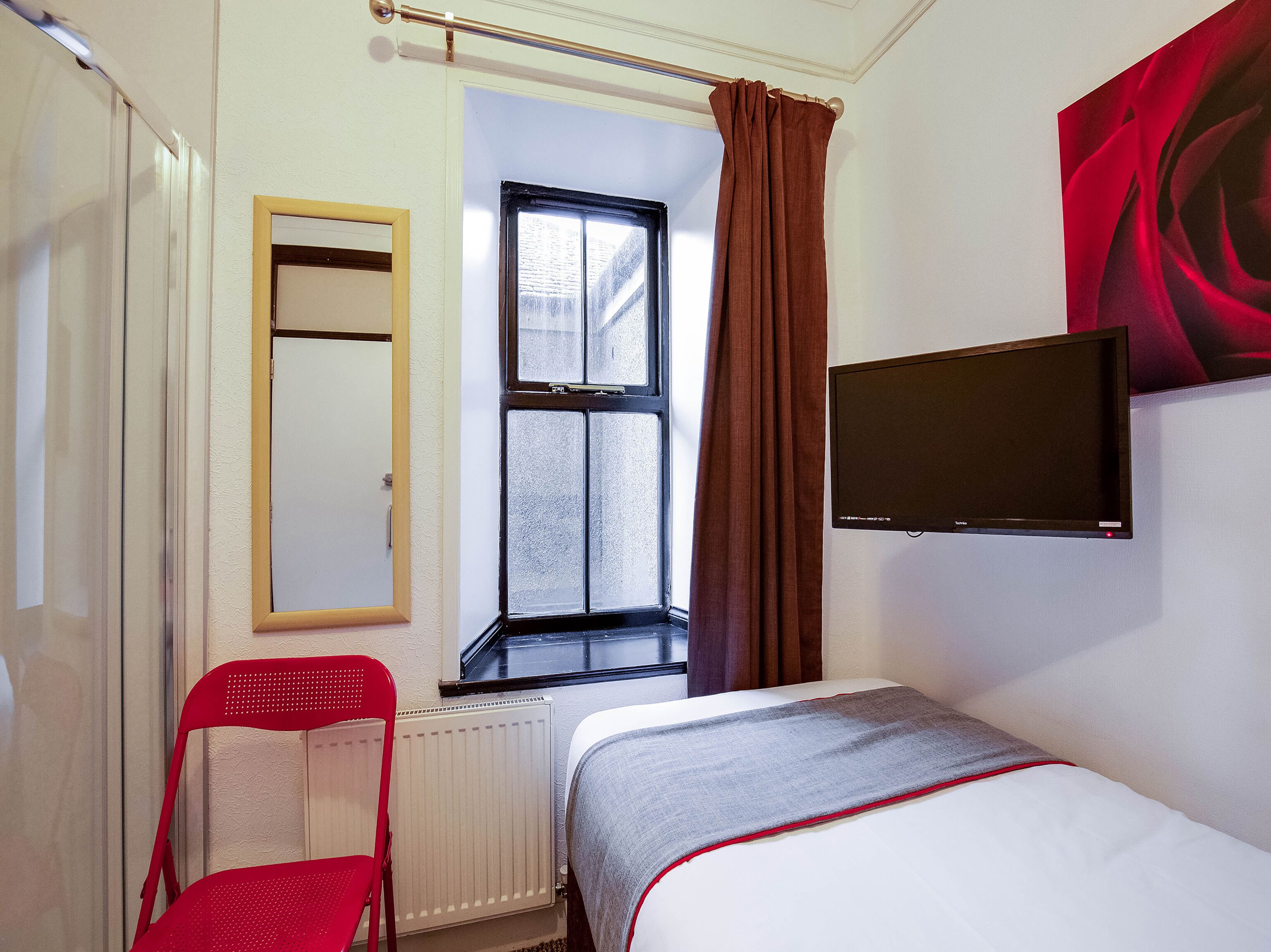 A hotel room with a bed, a red chair, a window with curtains, a mirror, and a TV on the wall.