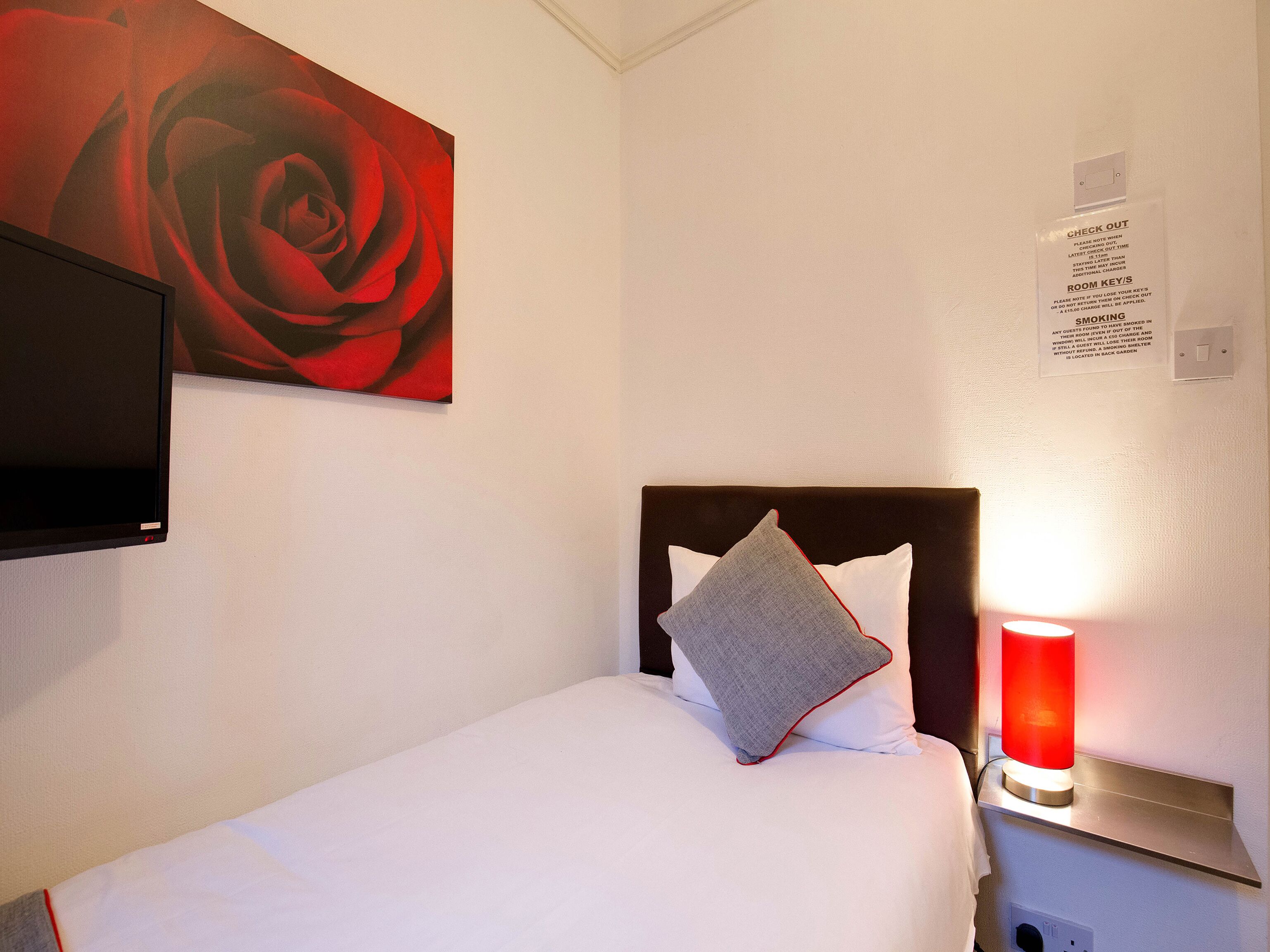 Una habitación de hotel con una cama, un cuadro de una rosa roja, un televisor, una mesita de noche con una lámpara roja y un cartel sobre llaves de la habitación y política sobre fumar.